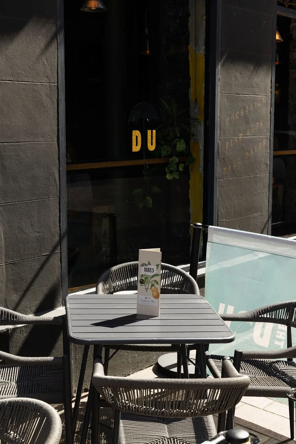 This image captures a sun-drenched outdoor seating area adjacent to a dark, urban building facade. A square slatted dark grey table is surrounded by modern woven rope chairs, with a menu featuring citrus illustrations standing prominently. The dark window reveals glimpses of interior greenery and bold gold lettering, while the exterior wall subtly advertises "COCKTAILS, FINE ALES, TALL TALES."