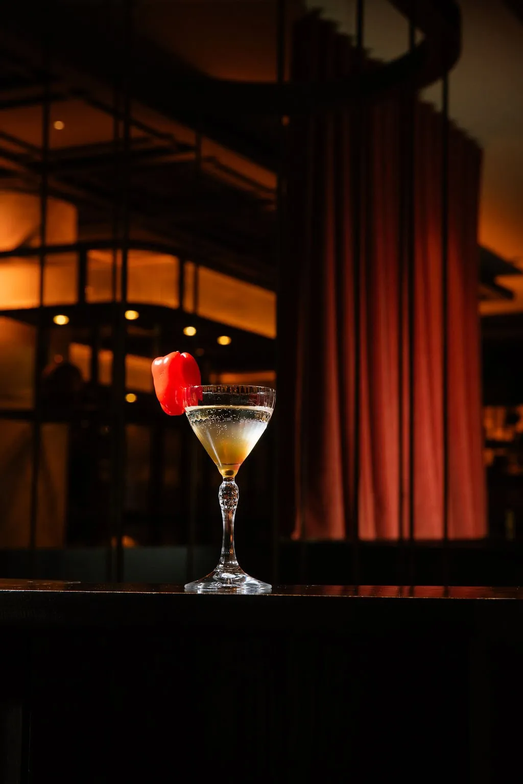 A sophisticated cocktail garnished with a vibrant red bell pepper sits on a dark bar, set against a warmly lit, blurred interior background featuring architectural details and a rich red curtain.