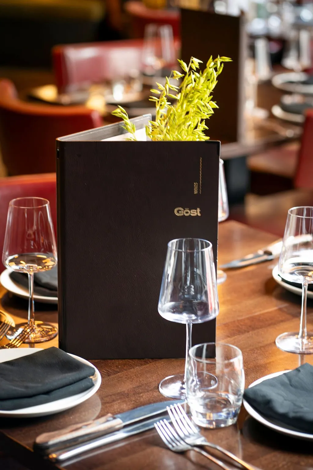 This close-up captures a meticulously set dining table within a sophisticated restaurant, featuring a dark stained wooden surface. A dark leather-bound menu, embossed with a gold "Göst" logo and holding a sprig of fresh green foliage, stands prominently amidst elegant stemware, white ceramic plates, and crisp black napkins. The warm, ambient lighting creates an intimate and inviting mood, with glimpses of rich red upholstered seating in the softly blurred background hinting at a luxurious dining experience.