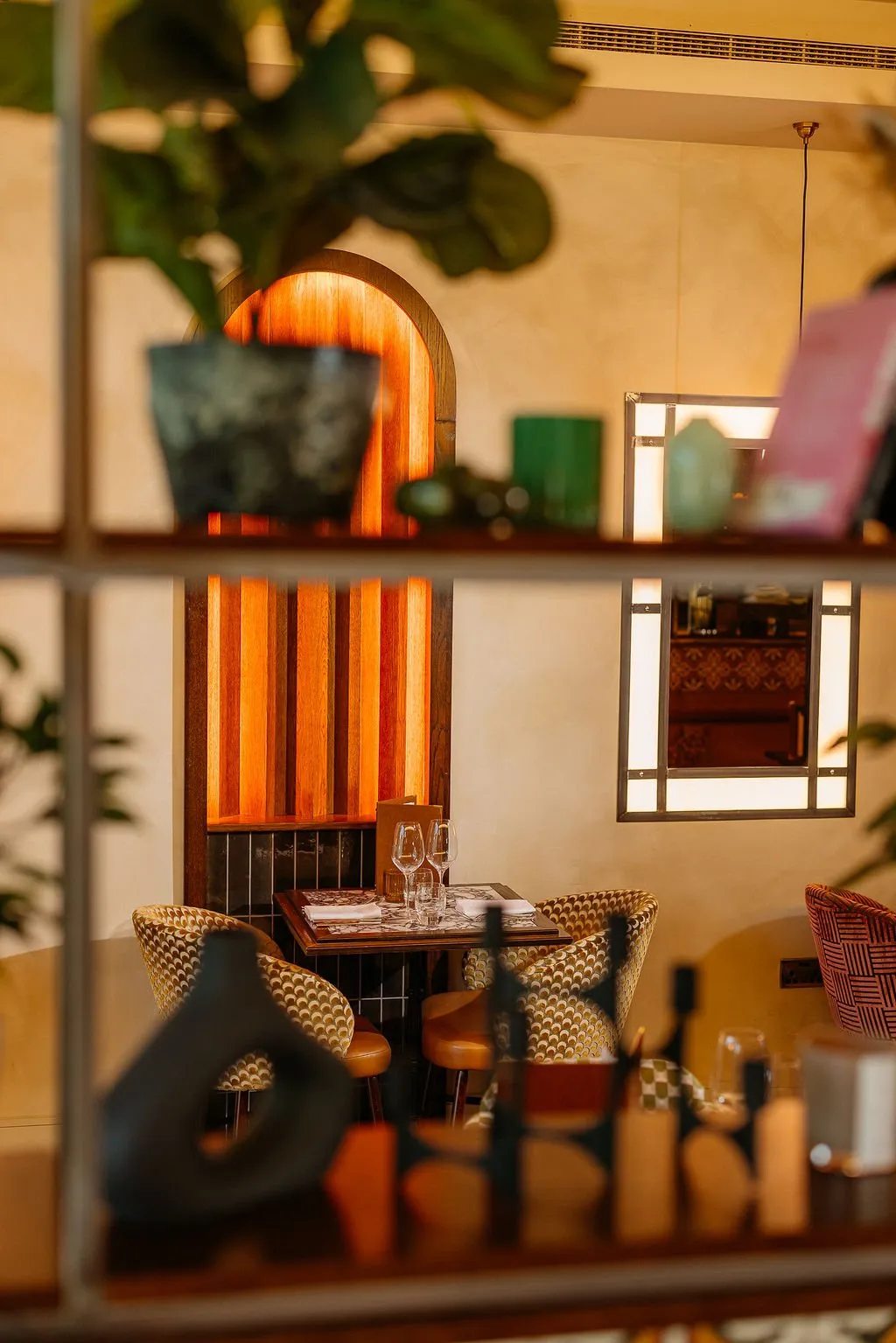 This intimate dining nook showcases a warm, sophisticated ambiance, perfect for a restaurant or bar. A striking backlit wooden archway feature with vertical slats creates a focal point, complemented by dark square tiles below. The space is furnished with a dark wood table set for two, flanked by chairs upholstered in a rich, geometric patterned fabric and accented with leather-look seats, all bathed in soft, inviting ambient light from the archway and an illuminated framed mirror.