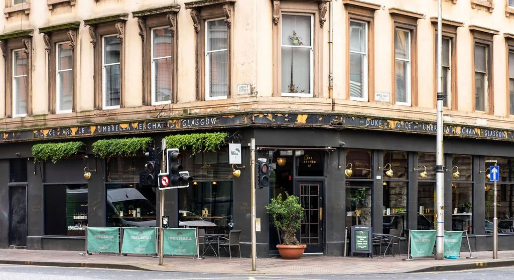 This street-level shot captures the charming exterior of a corner hospitality venue, featuring a dark, traditional facade with ornate gold lettering and peeling paint that hints at its history. Lush green planters adorn the top of the windows, complemented by classic brass wall sconces, creating an inviting glow. Outdoor seating with dark metal tables and chairs, framed by branded green barriers, offers a casual street-side experience, while glimpses through the large windows suggest a warm, intimate interior.
