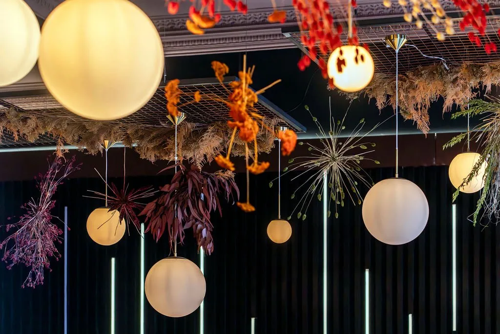 This close-up captures a dramatic and sophisticated overhead installation in a hospitality space, featuring an array of frosted spherical pendant lights with brass accents suspended amidst a rich tapestry of dried botanical arrangements. The warm, diffused glow from the pendants contrasts with cool linear LED strips on the dark, vertically paneled background, creating an intimate and modern ambiance. The organic shapes and autumnal hues of the botanicals, including ferns, grasses, and various seed pods, are artfully arranged on a metal grid, adding texture and a natural, yet refined, touch to the contemporary design. The overall effect is one of curated elegance and warmth, perfect for an upscale dining or lounge environment.