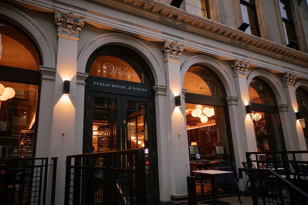 An inviting low-angle exterior shot of a historic building's ground floor, featuring arched windows and a 'Public House & Dining' entrance, warmly lit at dusk.