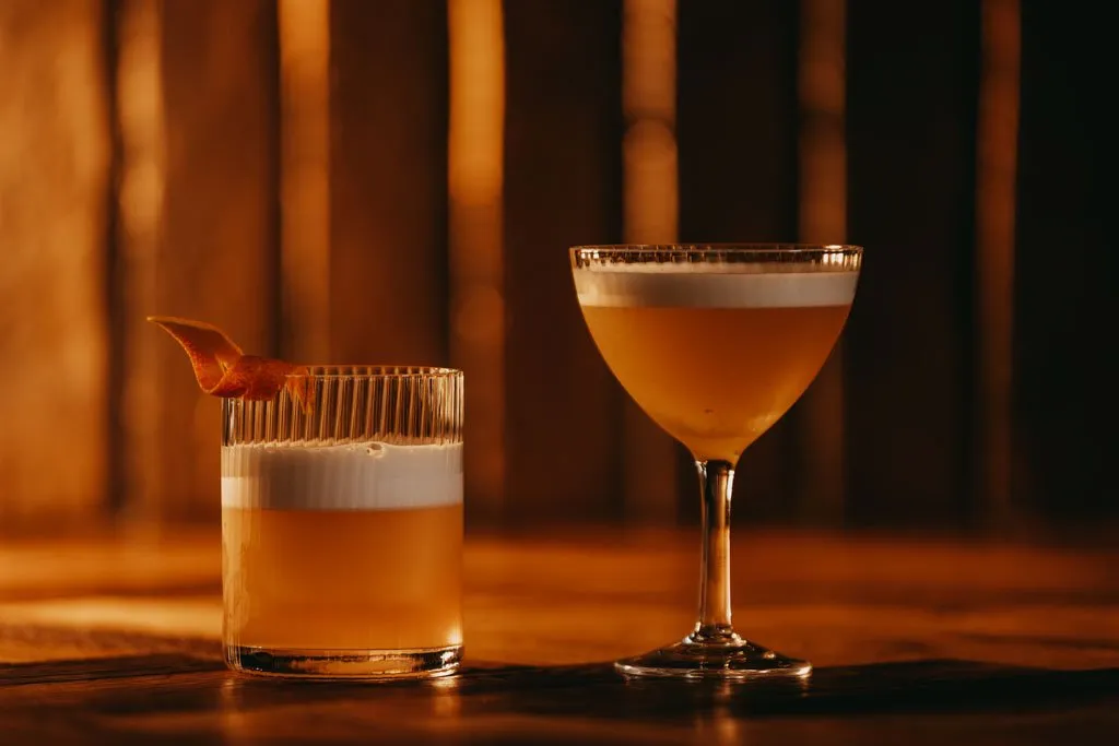 Two sophisticated cocktails are presented on a rich, dark wooden surface, bathed in warm, ambient lighting that casts soft reflections and highlights the intricate textures. The background features blurred vertical elements, likely wooden slats, creating a sense of depth and intimacy. One drink, in a fluted rocks glass with an elegant orange peel garnish, sits beside another in a classic coupe, both topped with a delicate foam, evoking a luxurious and inviting evening atmosphere.