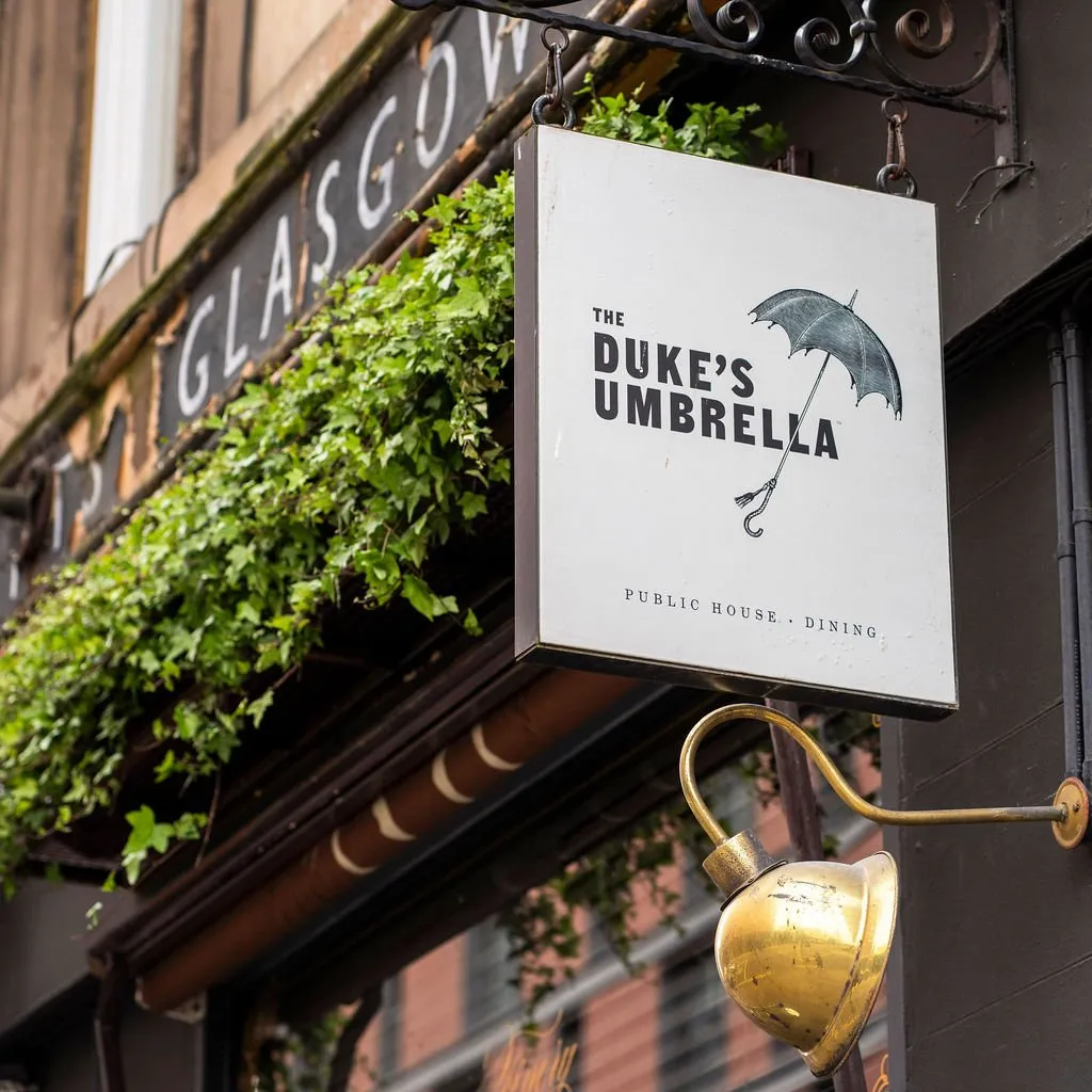 This close-up exterior shot showcases the charming facade of a traditional public house. A prominent white hanging sign for "The Duke's Umbrella" features a classic black umbrella graphic and bold typography, suspended by ornate black wrought ironwork. Below the sign, a lush green ivy planter spills over a dark brown window box, adding natural texture against the dark grey building facade. A polished brass gooseneck spotlight is mounted beneath the sign, poised to illuminate it, hinting at a warm, inviting atmosphere for evening patrons.