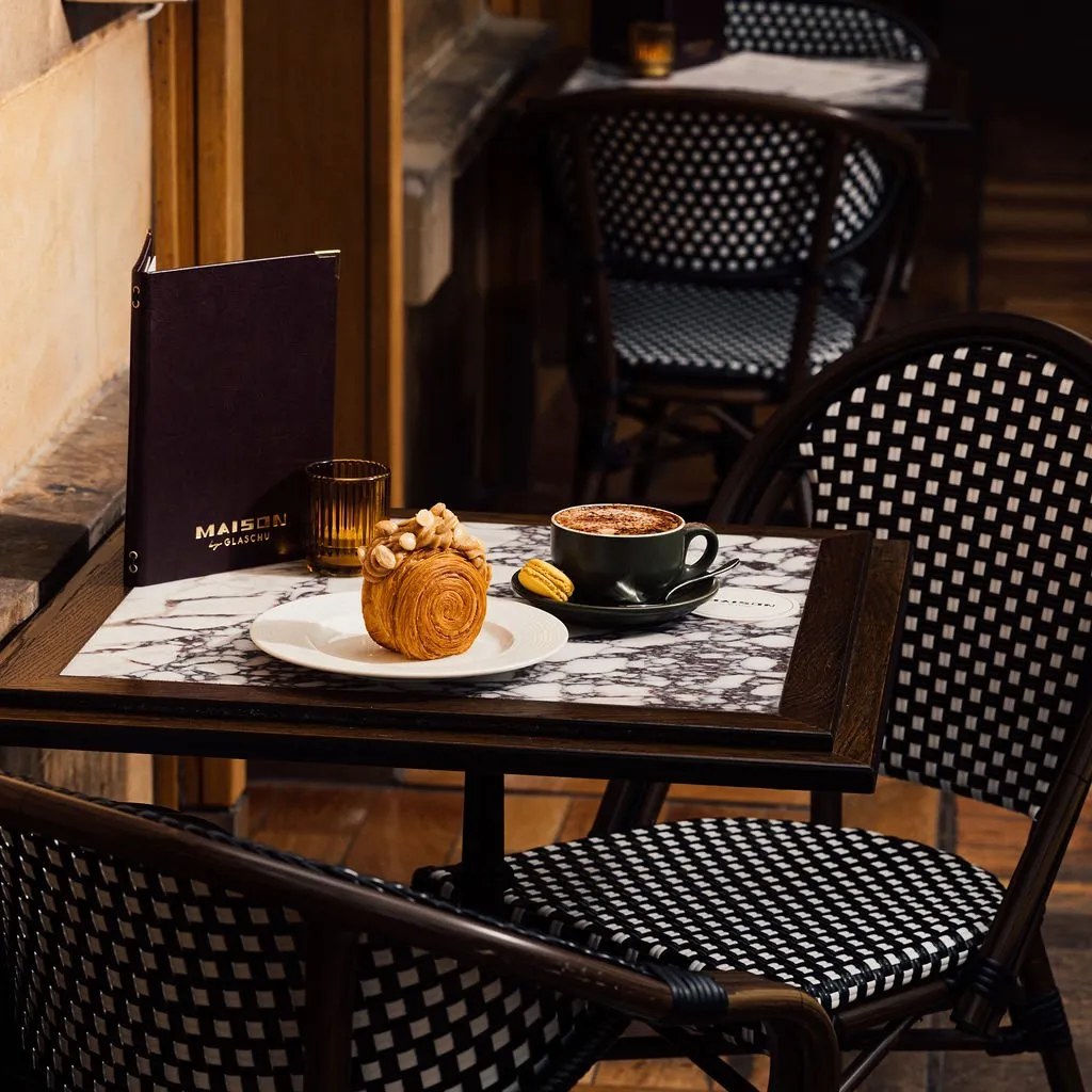 This intimate shot captures a charming cafe table setting, featuring a dark wood table with a white and grey veined marble top. Two classic bistro chairs, crafted from dark wood or rattan with distinctive black and white checkerboard woven seats and backs, flank the table. A delectable croissant, a dark ceramic cup of coffee, a small macaron, and a dark menu with gold lettering are artfully arranged, all bathed in warm, soft lighting that evokes a cozy, inviting atmosphere.