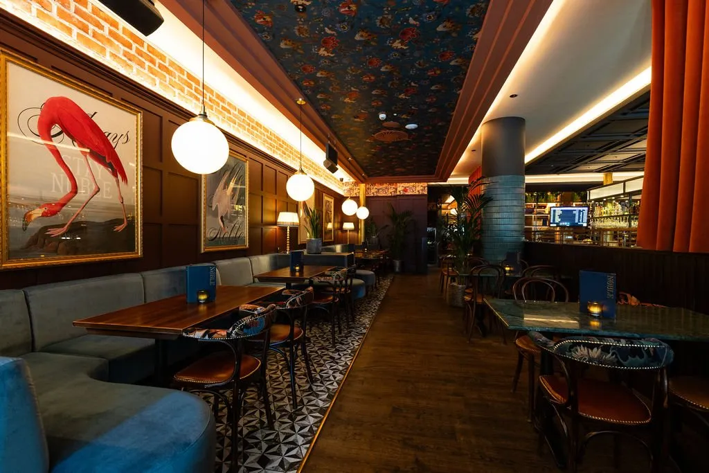 A vibrant and eclectic bar or restaurant interior features a long banquette, a richly patterned ceiling, and diverse seating arrangements under warm lighting.