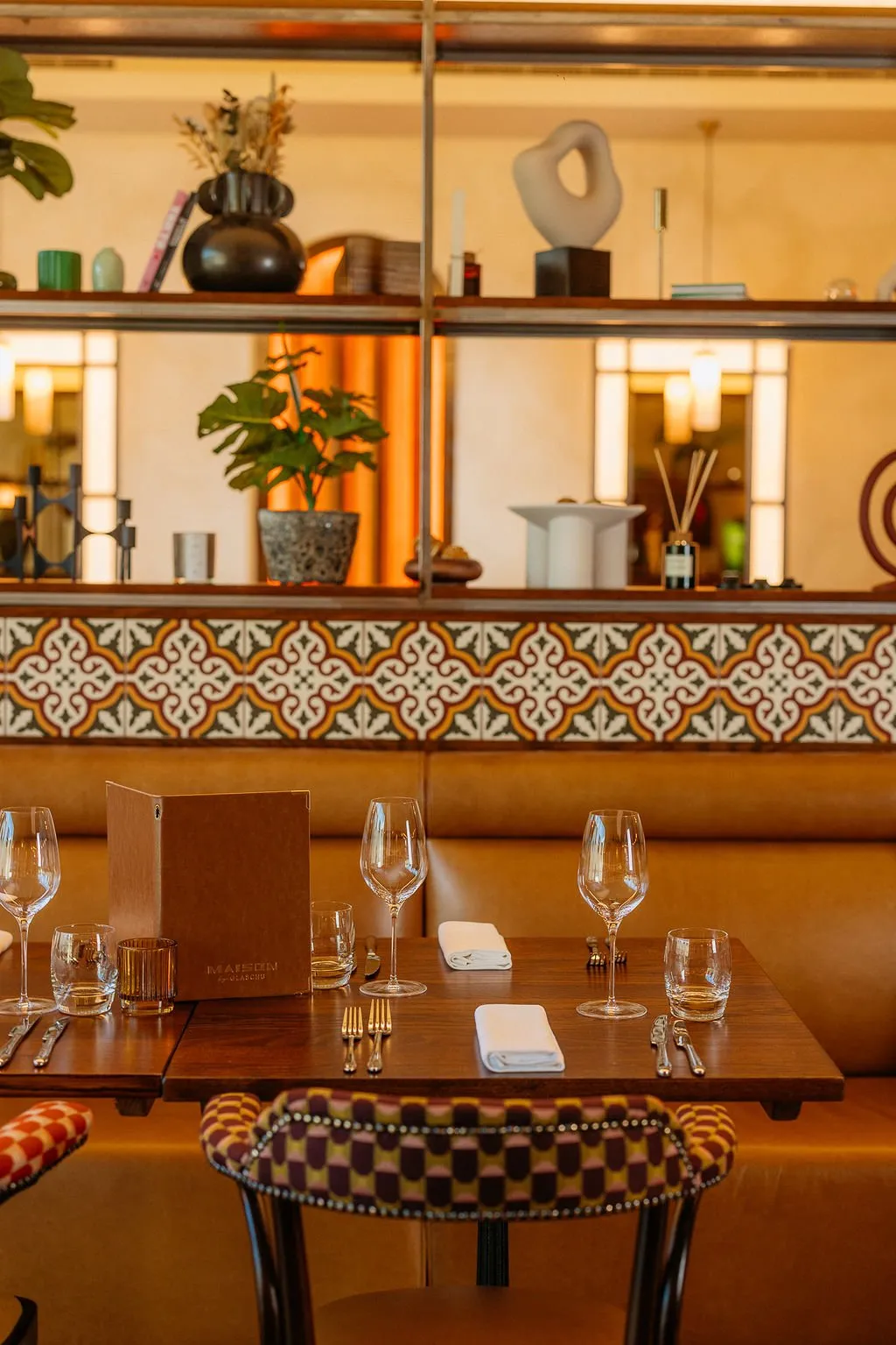 This inviting restaurant interior features a warm, sophisticated ambiance, centered around a dark wood dining table set for two against a plush tan leather banquette. Above the banquette, a striking band of patterned ceramic tiles adds a touch of Mediterranean flair, topped by a metal-framed shelving unit displaying curated decorative objects and lush greenery. The lighting is soft and ambient, creating a cozy and intimate dining experience, complemented by a vibrant, geometrically patterned dining chair.
