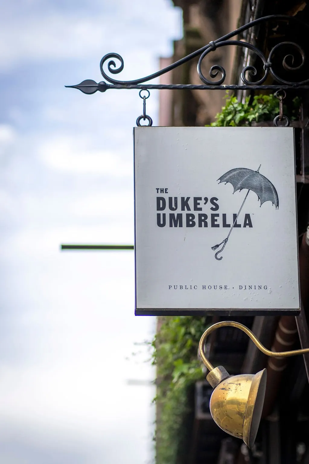 A detailed exterior shot captures the classic signage for "The Duke's Umbrella," a public house and dining establishment. The white rectangular sign, featuring a black umbrella graphic and elegant typography, hangs from an ornate, dark wrought iron bracket with decorative scrollwork. Below the sign, a vintage-style brass gooseneck light fixture is mounted, designed to illuminate the sign, while hints of green foliage and a building facade are visible in the soft daylight background.