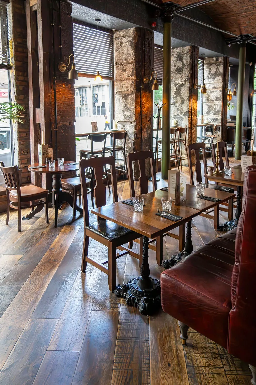 This inviting hospitality space features a rich, industrial-rustic aesthetic, characterized by exposed brickwork, rough-hewn timber columns, and distressed plaster walls. Warm brass sconces and industrial pendants cast a cozy glow over dark wood tables with ornate cast iron bases, complemented by classic wooden chairs with leather seats and a prominent deep red leather banquette. The wide plank distressed wood flooring and large street-facing windows with blinds complete the warm, lived-in atmosphere, perfect for a relaxed dining or drinking experience.