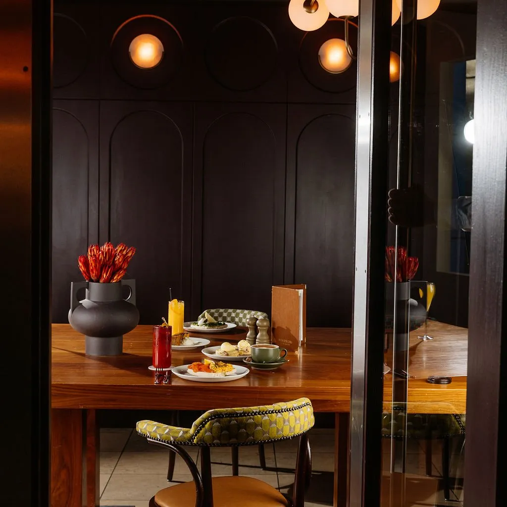 This intimate dining scene features a rich, dark aesthetic, centered around a polished wooden table set for a meal. The backdrop is a sophisticated dark paneled wall with subtle arched details, illuminated by warm, circular wall sconces that cast a soft, inviting glow. Upholstered chairs with a distinctive geometric pattern in olive green and a warm leather seat add a touch of vintage elegance, complementing the vibrant protea arrangement and various dishes on the table, all framed through a dark doorway.