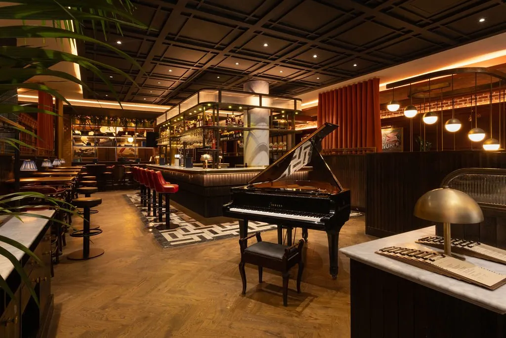 A sophisticated and warmly lit bar and lounge area featuring a grand piano, a well-stocked bar, and various seating options under a geometric ceiling.