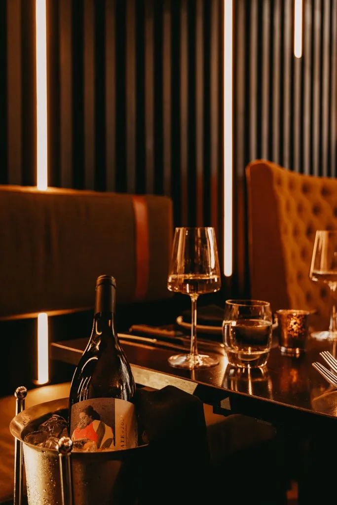This intimate restaurant or bar scene features plush mustard velvet banquette seating, one with classic tufting, set against a dark, vertically slatted wall. Warm, linear LED strip lighting punctuates the background, casting a soft, inviting glow over the dark, reflective tabletop. A chilled bottle of wine in a silver ice bucket, elegant stemware, and a flickering votive candle create a luxurious and cozy dining atmosphere.