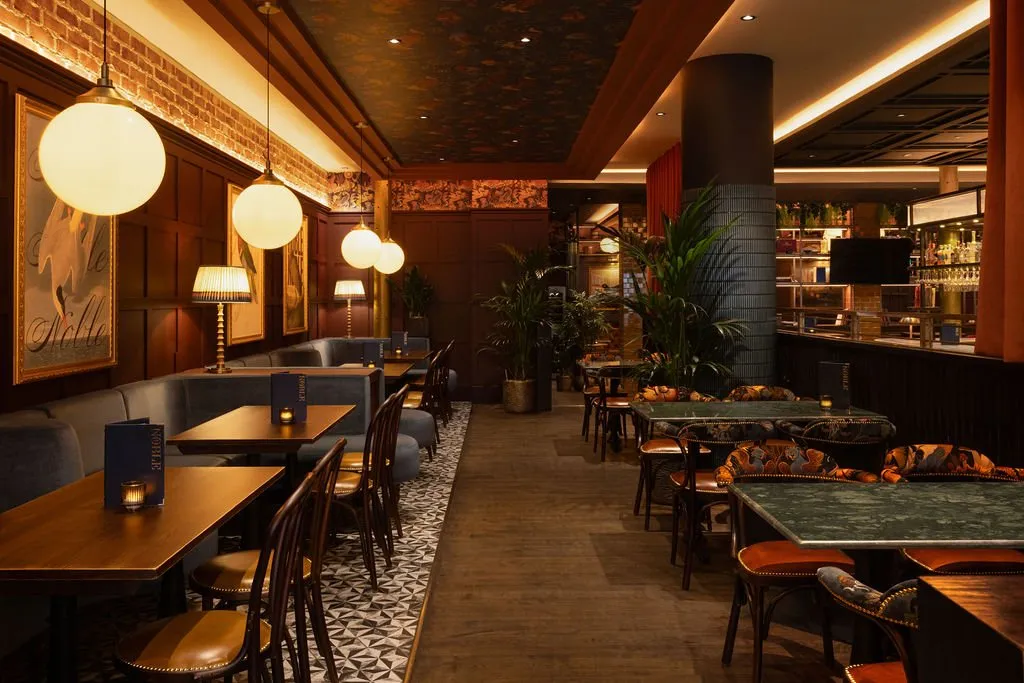 A sophisticated and warmly lit restaurant or bar interior featuring banquette seating, dark wood paneling, patterned elements, and a well-stocked bar in the background.