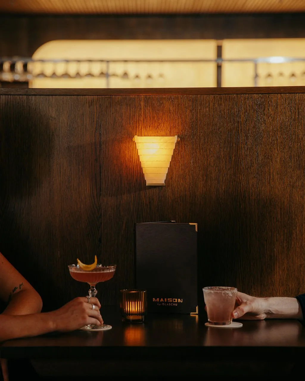 This intimate shot captures a cozy corner within a sophisticated bar or restaurant, featuring a dark stained wood partition that grounds the scene. A striking tiered wall sconce emits a warm, inviting glow, complemented by a flickering candle on the dark tabletop where two patrons enjoy meticulously crafted cocktails. The rich wood tones and soft ambient lighting create an atmosphere of refined comfort, perfect for an evening gathering.