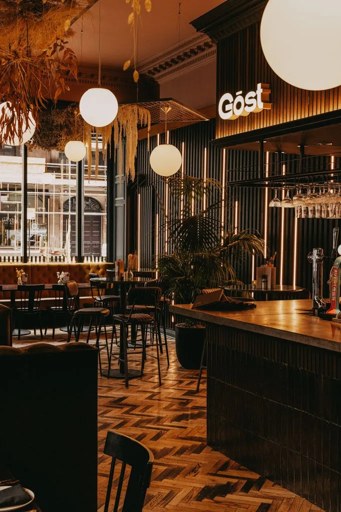 This sophisticated bar and restaurant interior features a warm, inviting ambiance, illuminated by a mix of spherical pendant lights and dramatic vertical LED strips integrated into dark fluted wall panels. A long, dark-tiled bar with a light stone top anchors the space, complemented by a mix of plush velvet banquette seating and high-top tables with industrial-style stools. The design is enriched by a striking herringbone wood floor and abundant dried floral installations suspended from the high ceilings, creating a modern yet organic feel.