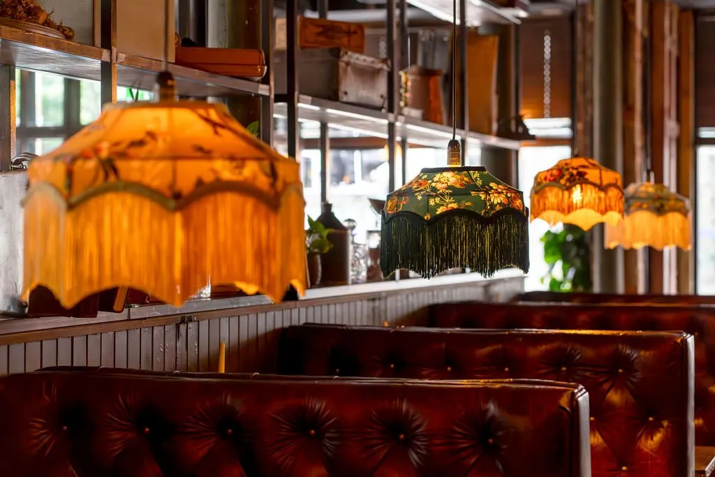 This intimate interior scene showcases a series of deep red-brown tufted leather booths, creating a cozy and inviting atmosphere. Overhead, an array of eclectic pendant lamps with fringed fabric shades, some featuring vibrant floral patterns, cast a warm, golden glow across the space. The background reveals dark wood wainscoting and metal shelving adorned with various decorative objects, hinting at a richly layered and vintage-inspired design.