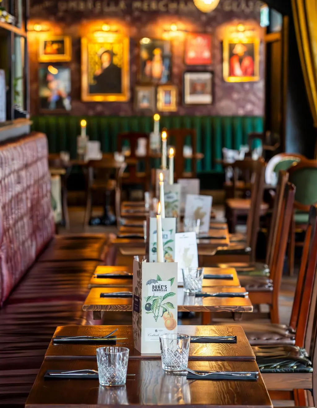 This interior shot captures a warm, inviting dining space, likely a restaurant or pub, with a long perspective down a row of dark wooden tables set for service. The background features a richly textured wall with dark patterned wallpaper and a deep green fluted banquette, adorned with an eclectic gallery wall of framed art illuminated by individual picture lights. Candlelight from the tables, combined with the warm wall lighting, creates an intimate and sophisticated atmosphere, highlighting the traditional yet refined aesthetic.