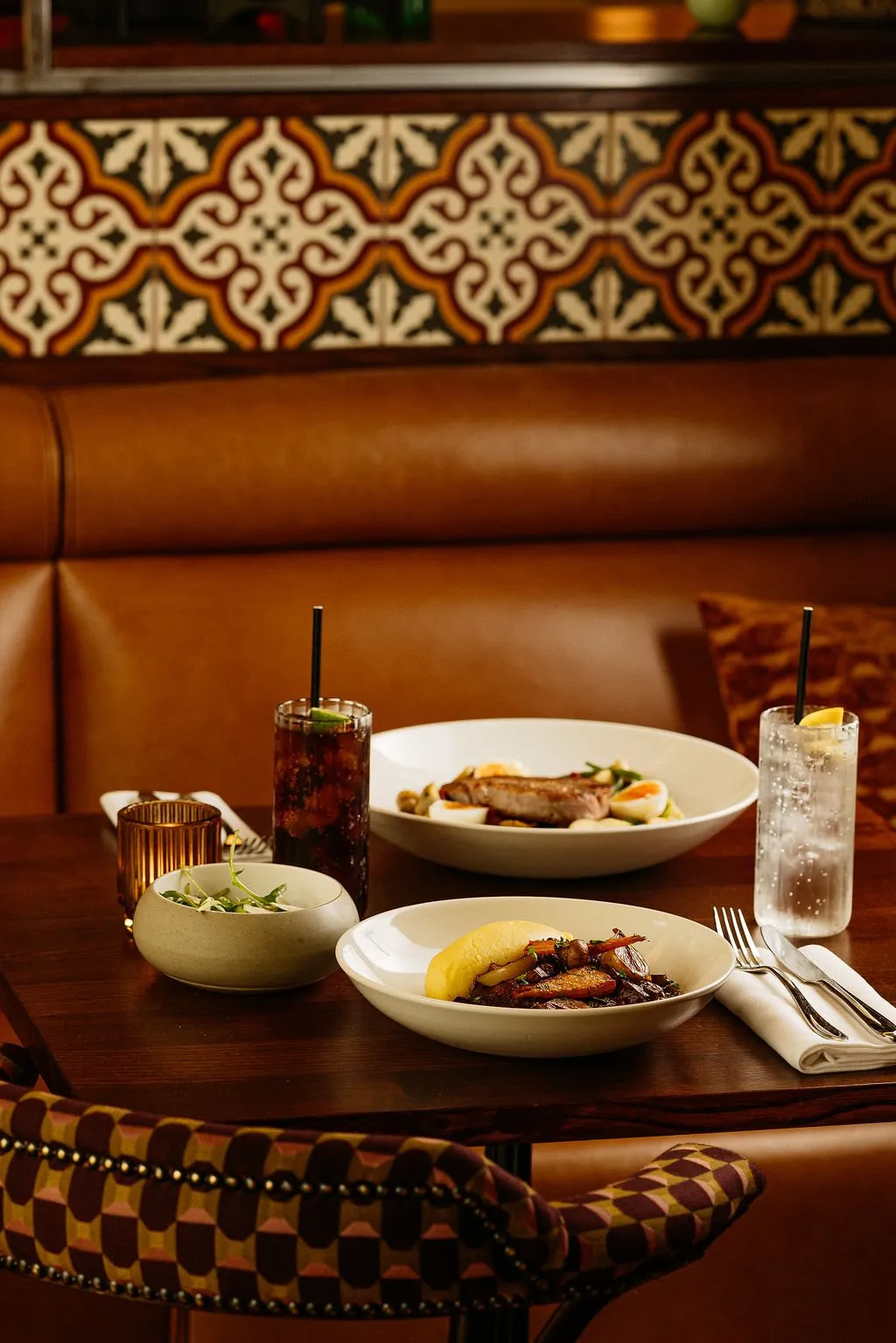This intimate dining scene features a dark polished wooden table set with modern ceramic dinnerware and fluted glass tumblers, offering a cozy spot for a meal. Behind the table, a plush, tufted caramel leather banquette provides comfortable seating, complemented by a striking wall panel of ornate, geometric patterned tiles in warm earthy tones. The foreground reveals a dining chair upholstered in a bold, geometric patterned fabric with studded trim, all bathed in warm, inviting ambient lighting that creates a sophisticated and relaxed atmosphere.