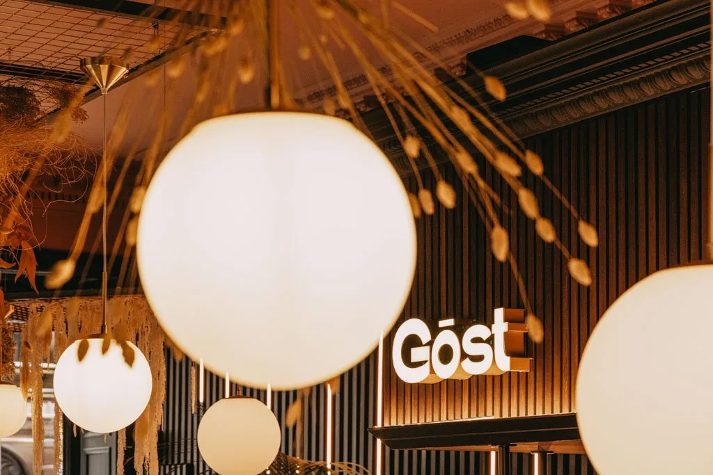 This close-up interior shot captures a warm and inviting atmosphere, dominated by a cluster of spherical, frosted glass pendant lights with brass accents hanging from an exposed grid ceiling. Behind them, a dark fluted wooden wall features an illuminated "Göst" sign, subtly backlit to enhance its presence. Dried, organic botanical elements are artfully integrated into the ceiling decor, adding texture and a natural touch to the sophisticated, dimly lit space, suggesting an intimate dining or lounge setting.