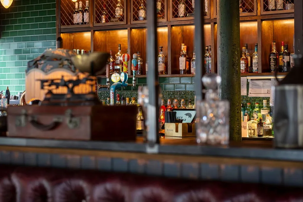 This inviting bar interior features a rich material palette and warm, atmospheric lighting. The back bar is clad in glossy emerald green subway tiles, contrasting with dark wooden shelving units that display an array of spirits, each shelf warmly lit from within. A deep red, tufted leather banquette or bar stool base is visible in the blurred foreground, hinting at comfortable seating. Decorative elements like an antique-style suitcase and crystal decanters add a touch of vintage charm, while industrial-style metalwork frames the scene.