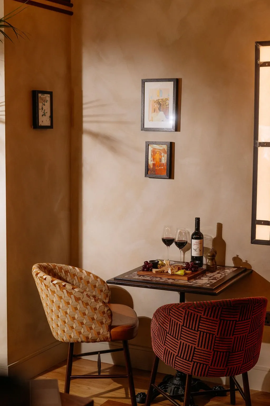 This intimate corner setting features a small square table with a dark frame and a striking marble-patterned top, flanked by two distinct upholstered chairs. The walls are finished in a warm, textured plaster, adorned with framed artwork and illuminated by a soft, directional light that casts intriguing shadows, including a palm frond. The chairs, one in a geometric gold-and-brown pattern and the other in a terracotta linear weave, add an eclectic yet sophisticated touch, creating a cozy and inviting atmosphere perfect for a quiet evening.