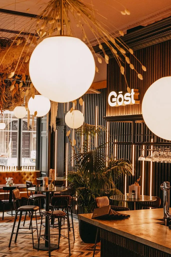 This inviting hospitality interior features a sophisticated blend of textures and warm lighting, creating a cozy yet modern atmosphere. Dark fluted wall panels are dramatically lit by integrated vertical LED strips and a prominent "Göst" neon sign, contrasting with a warm herringbone wood floor and a concrete-look bar top. The space offers diverse seating, including a plush mustard velvet banquette by large windows and high-top tables with ribbed upholstered bar stools, all illuminated by oversized spherical pendant lights and striking dried botanical ceiling installations. Lush green plants add a touch of natural vibrancy to the scene.