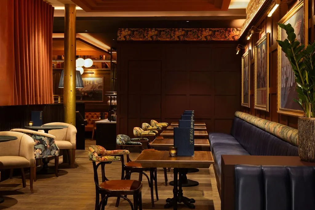 A luxurious and richly decorated restaurant or bar interior features a mix of banquette seating, upholstered chairs, and dark wood tables under warm, ambient lighting.