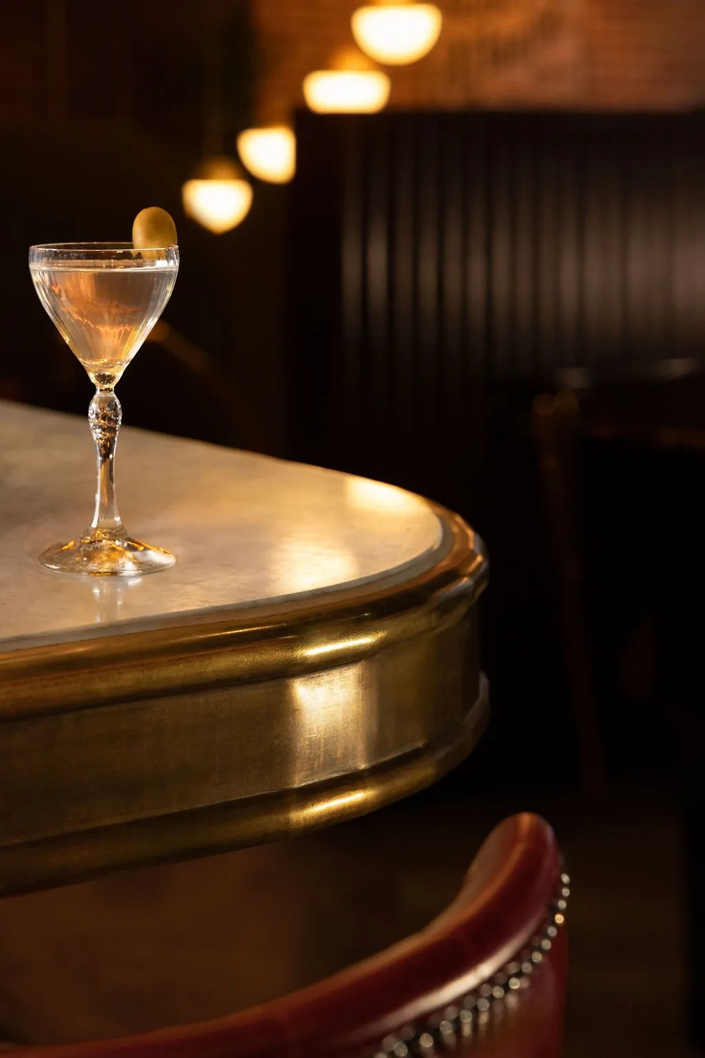 A close-up shot of a classic martini with an olive, resting on a polished marble and brass bar counter in a dimly lit, sophisticated interior.