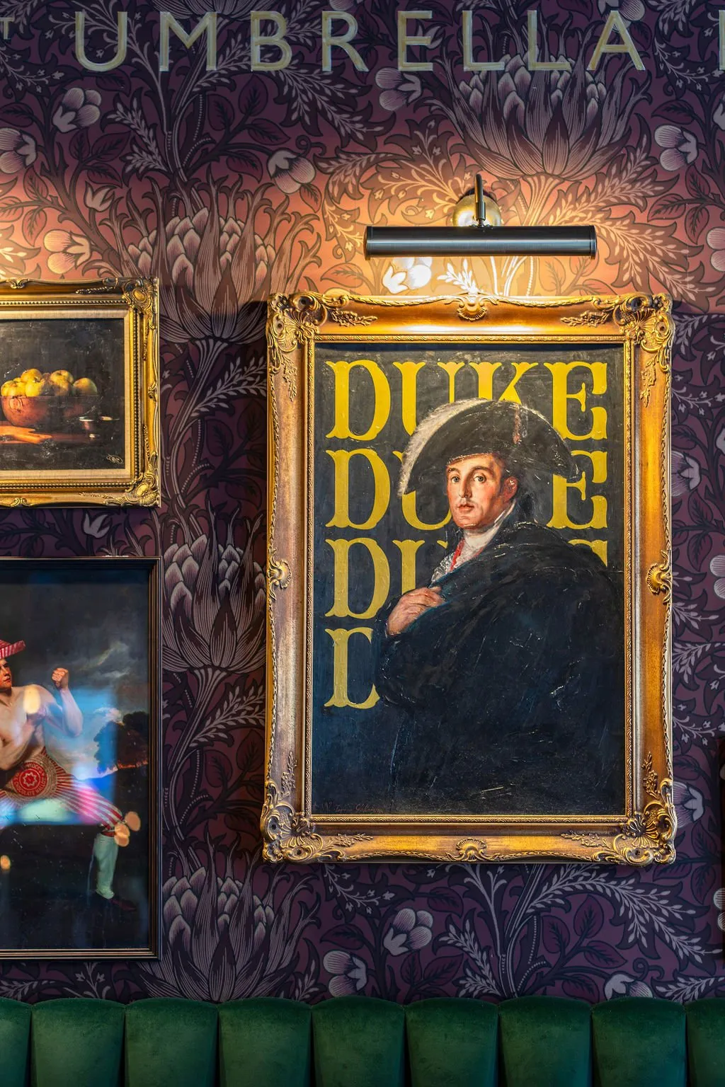 This close-up shot reveals a richly textured wall featuring a deep plum-toned, intricate floral wallpaper that subtly integrates the "UMBRELLA" brand name at the top. A collection of framed artworks, including a prominent portrait of a man with "DUKE" emblazoned in yellow, are dramatically lit by a sleek black picture light, casting a warm glow. Below, the plush, deep emerald green velvet upholstery of a banquette hints at a luxurious and inviting seating area, creating an atmosphere of classic sophistication with an eclectic twist.