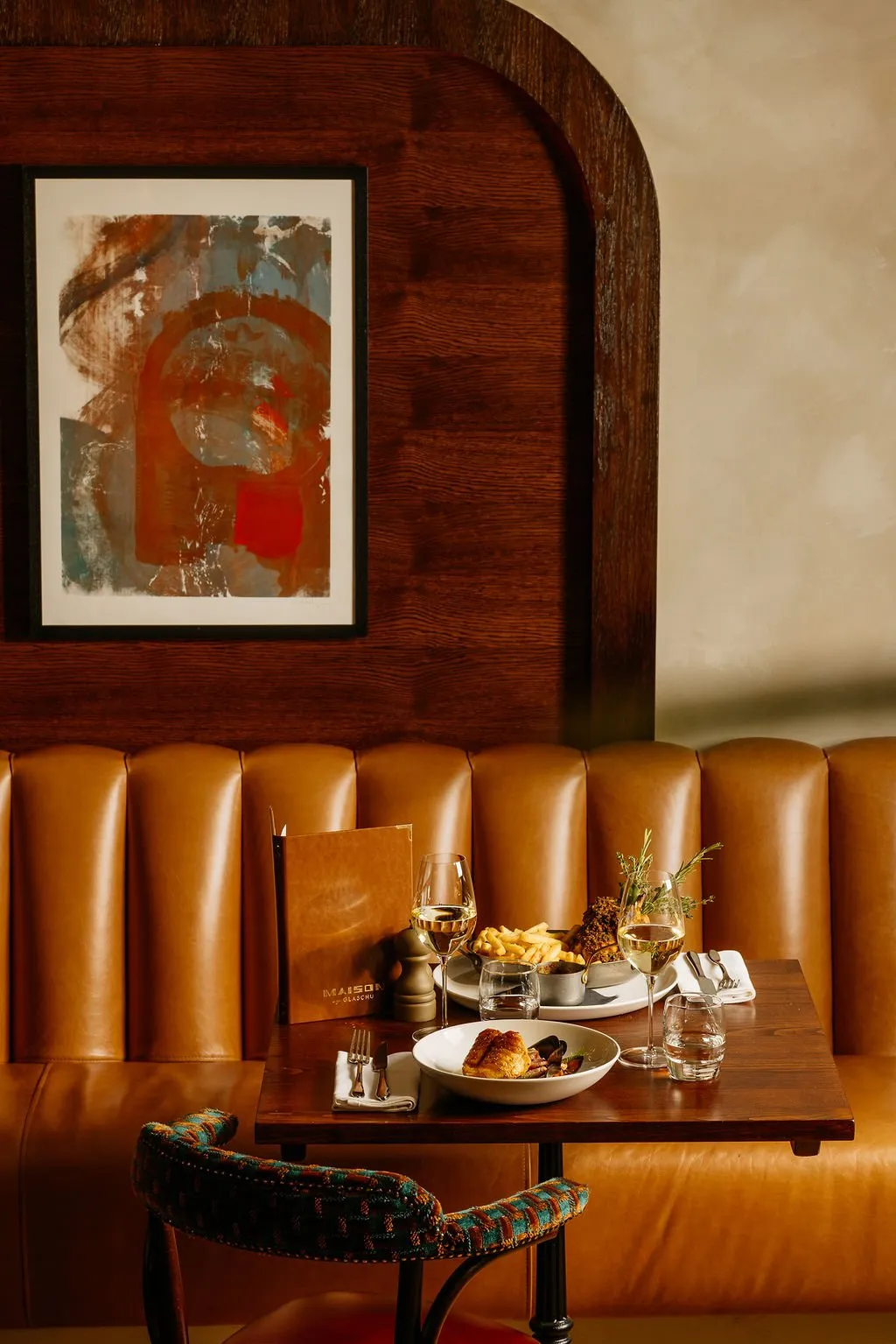This intimate restaurant booth features a rich, dark stained oak paneling with a distinctive arched detail, framing an abstract artwork in warm tones. Below, a plush, tan leather fluted banquette provides comfortable seating, complemented by a dark wood table set for two with a vibrantly patterned upholstered chair. The warm, dim lighting creates a sophisticated and inviting atmosphere, highlighting the classic yet eclectic material palette.