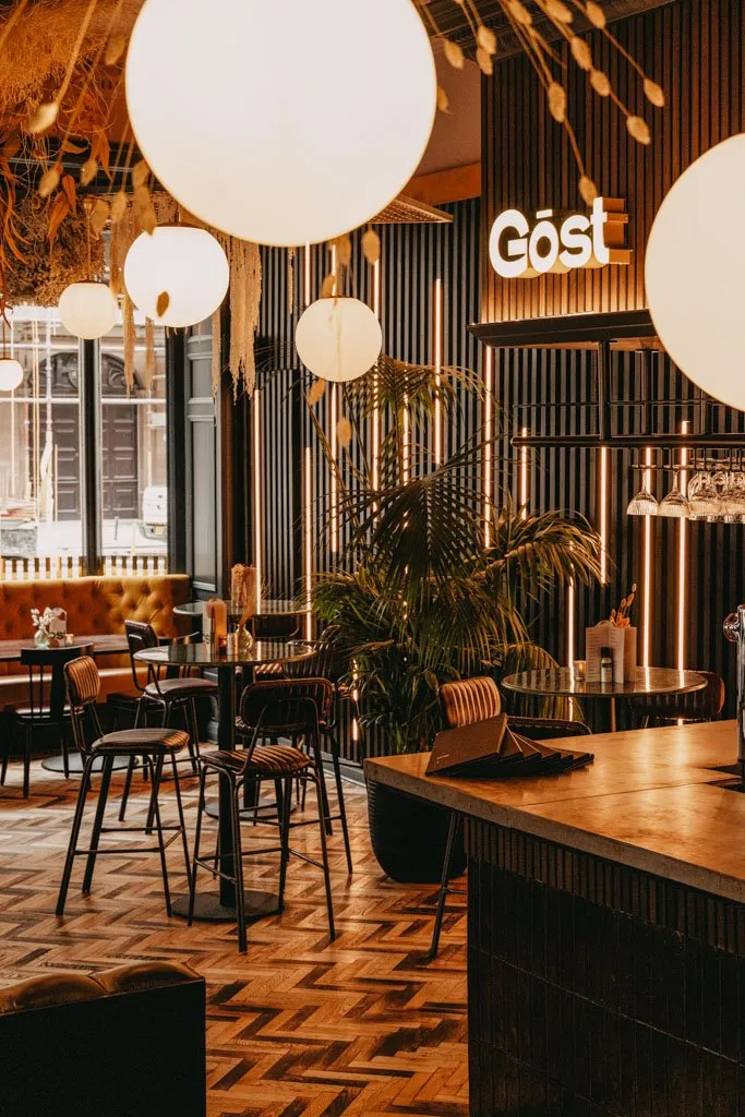 This inviting restaurant interior features a rich material palette and layered lighting. A tufted mustard velvet banquette lines the window, complemented by dark metal tables and chairs with ribbed upholstery, all set against a striking dark slatted wall with integrated warm amber LED strips. Overhead, large spherical pendant lights and dried botanical installations create a soft glow, while a herringbone wood floor and a textured bar counter complete the sophisticated, cozy atmosphere.