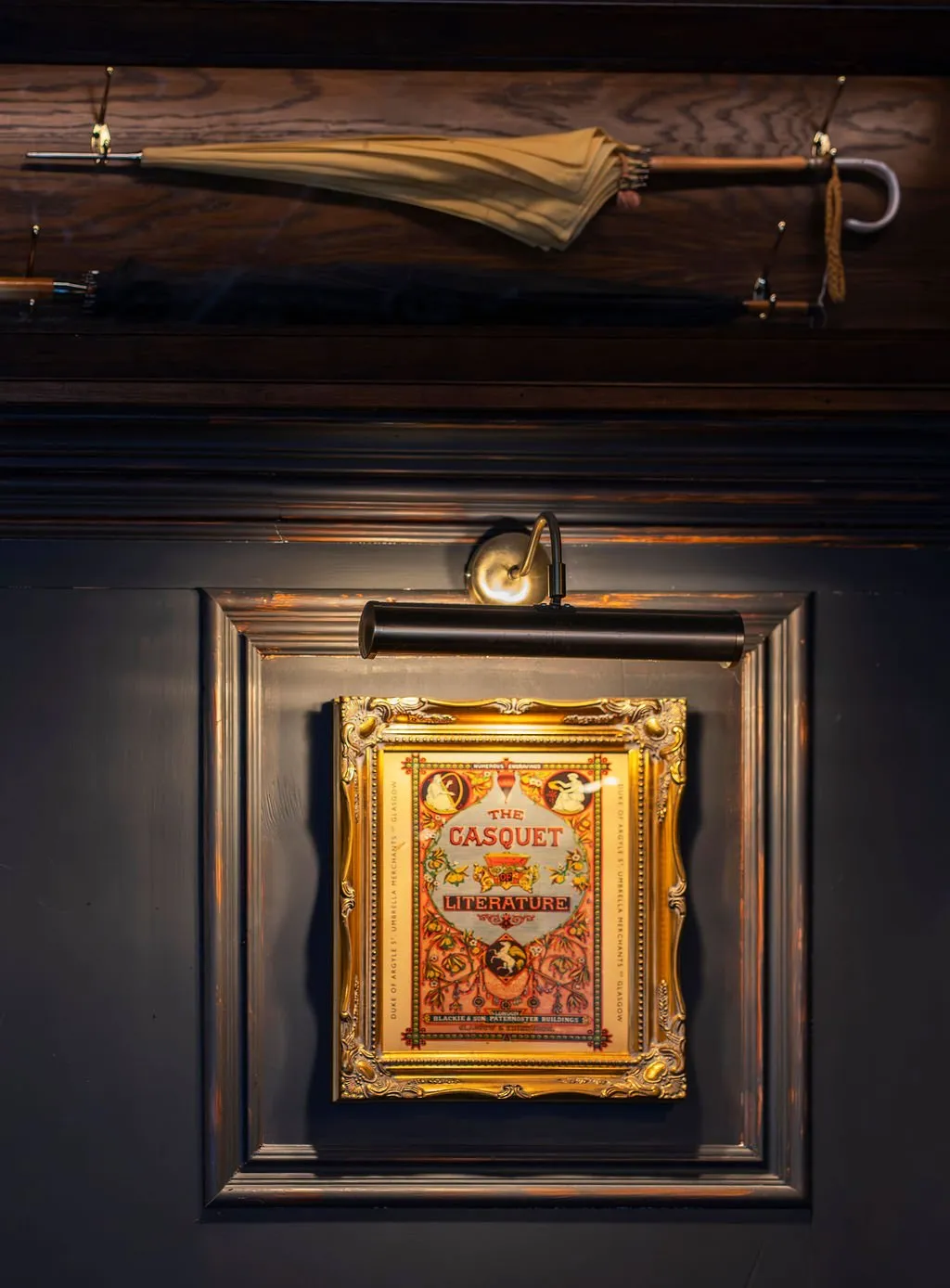 This close-up shot reveals a sophisticated, dimly lit interior featuring dark, richly paneled walls. A vintage-style print titled "The Casquet Literature," encased in an ornate gold frame, is prominently displayed and illuminated by a sleek brass and black picture light. Above, a dark wooden shelf or panel holds two classic umbrellas on elegant brass hooks, contributing to a refined, club-like atmosphere with a strong sense of heritage.