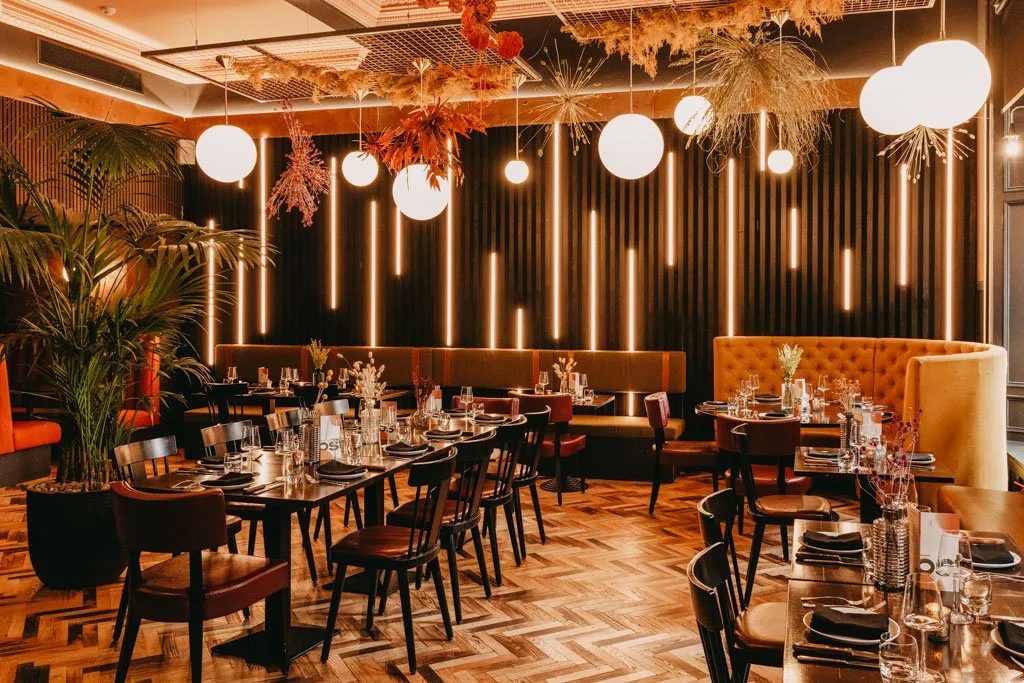 This vibrant restaurant interior features a striking dark slatted feature wall illuminated by vertical warm LED strips, creating a dynamic backdrop. Spherical pendant lights and an elaborate dried botanical ceiling installation add layers of decorative lighting and texture. The space is furnished with a mix of dark wood dining tables and chairs, complemented by rich orange and olive-toned upholstered banquette and booth seating, all set upon a warm herringbone wood floor. The overall mood is intimate and sophisticated, enhanced by the warm ambient lighting and lush potted plants.