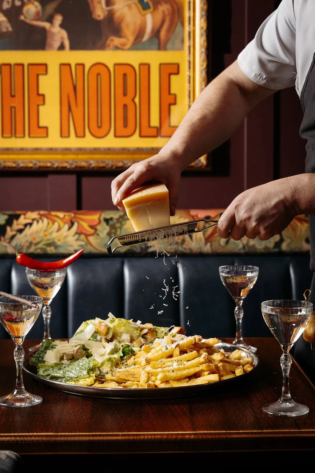 A close-up shot captures a chef grating cheese over a plate of fries and Caesar salad, served alongside cocktails on a dark wooden table in a restaurant with vintage-inspired decor.