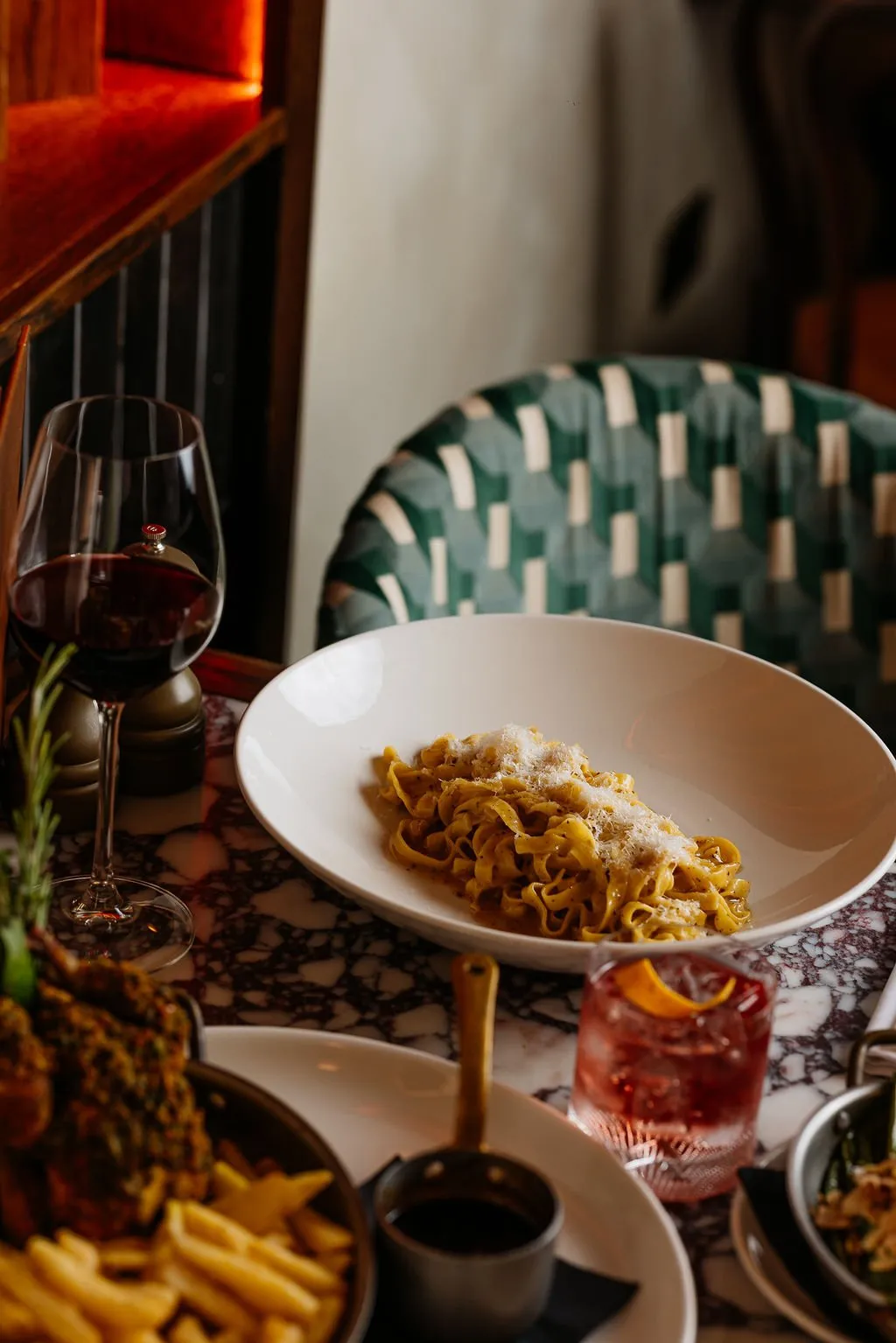 This close-up shot captures an intimate dining experience within a sophisticated restaurant setting. A dark, richly veined marble tabletop is laden with elegantly presented dishes, including pasta and a cocktail, alongside a glass of red wine. In the background, a curved dining chair upholstered in a striking dark green and cream geometric pattern adds a touch of Art Deco flair, complemented by the warm glow from unseen accent lighting on dark wood paneling, creating a cozy and luxurious evening atmosphere.