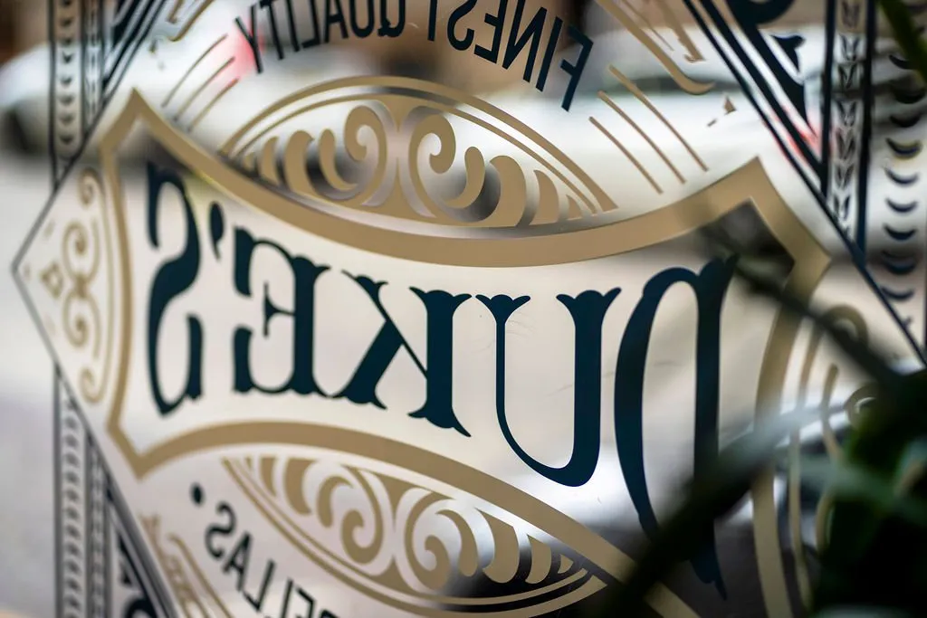 This detail shot captures an elegant, reverse-painted or etched glass sign, likely from a storefront or interior partition. The primary text "DUKE'S" is rendered in a sophisticated dark teal serif font, framed by intricate metallic gold scrollwork and decorative borders that evoke a classic, heritage aesthetic. The reflective quality of the glass, combined with the subtle ambient lighting, creates a sense of depth and refined craftsmanship, hinting at a venue with a strong brand identity and attention to traditional detailing.