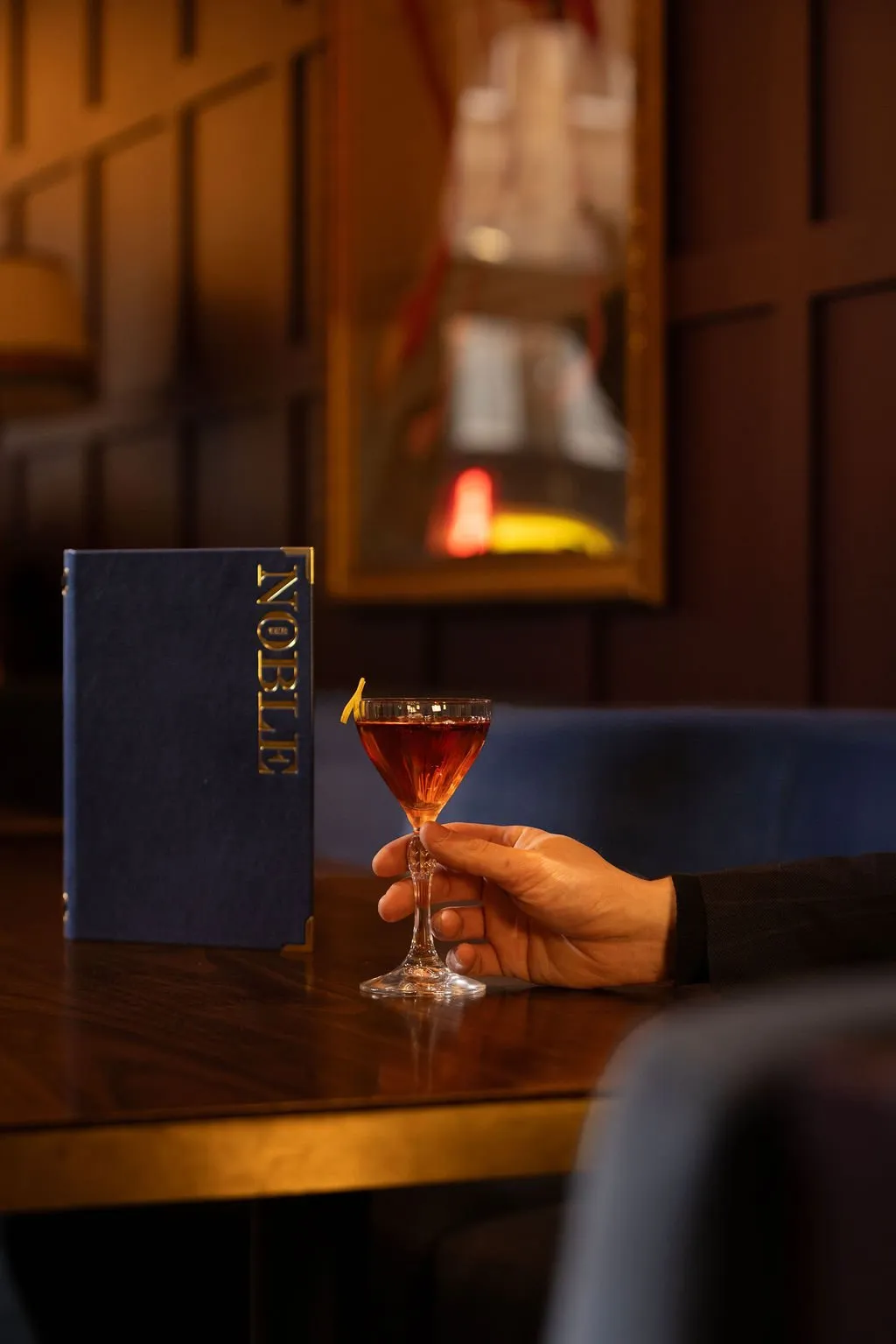 A close-up shot features a hand holding an amber cocktail with a lemon twist, positioned next to a dark blue menu with gold 'NOBLE' branding on a polished wooden table in a dimly lit, sophisticated bar.