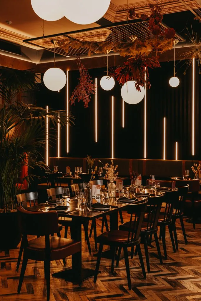 This moody and sophisticated restaurant interior features a dark palette illuminated by warm, ambient lighting. A mix of freestanding dark wood tables and chairs with rich leather seats are arranged alongside a deep upholstered banquette, all set upon a striking herringbone wood floor. Overhead, spherical pendant lights and a grid ceiling adorned with dried botanicals complement vertical LED strips on the dark fluted wall, creating an intimate and dramatic atmosphere. Ornate ceiling molding adds a touch of classic elegance to the contemporary design.
