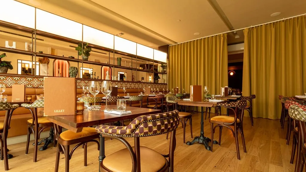 This inviting restaurant dining room features a warm, sophisticated palette with a mix of banquette and individual table seating. A long tan leather banquette runs along one wall, backed by intricate geometric tiles and an open shelving unit displaying decorative objects and plants, subtly lit by arched niches. The opposite side offers dark wood tables with cast iron bases, paired with classic bentwood chairs featuring mustard velvet seats and striking geometric patterned backrests, all under the soft glow of diffused overhead lighting and rich mustard-gold curtains.