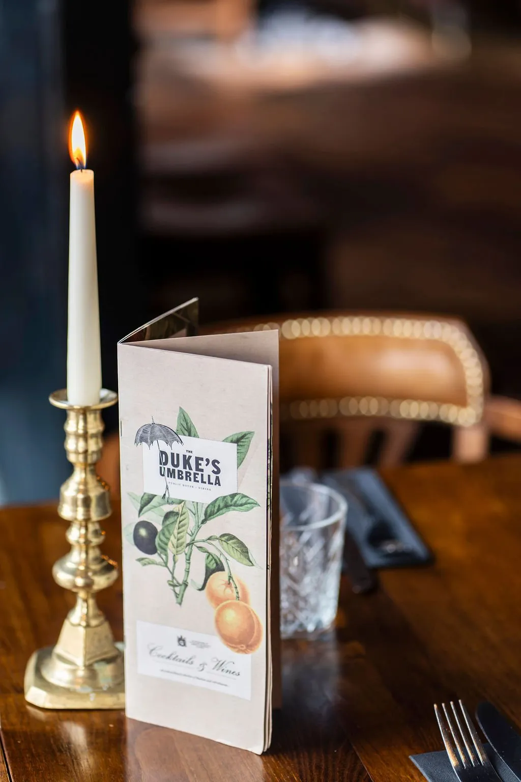 This intimate detail shot captures a beautifully set table in a dimly lit hospitality space, likely a pub or restaurant, creating a warm and inviting atmosphere. A slender, lit taper candle in a classic brass holder casts a soft glow, illuminating a bespoke menu featuring botanical illustrations of olives and oranges for "Duke's Umbrella." The dark wooden tabletop, paired with a cut-glass tumbler and sleek black cutlery, suggests a blend of traditional charm and contemporary refinement, with a blurred leather banquette in the background hinting at comfortable, classic seating.