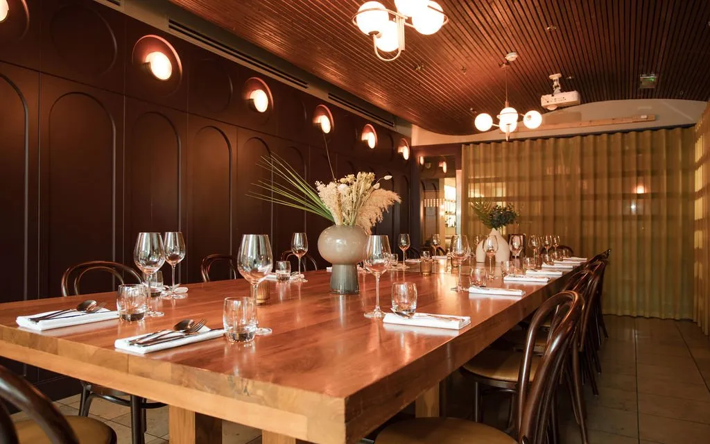 This intimate private dining room features a rich, warm palette, anchored by a long, solid timber communal table set for a formal meal. Dark arched wall paneling with circular sconces creates a sophisticated backdrop, complemented by a slatted timber ceiling and warm globe pendant lights. A translucent, wavy golden screen provides a soft partition, enhancing the cozy and elegant atmosphere, perfect for private events.