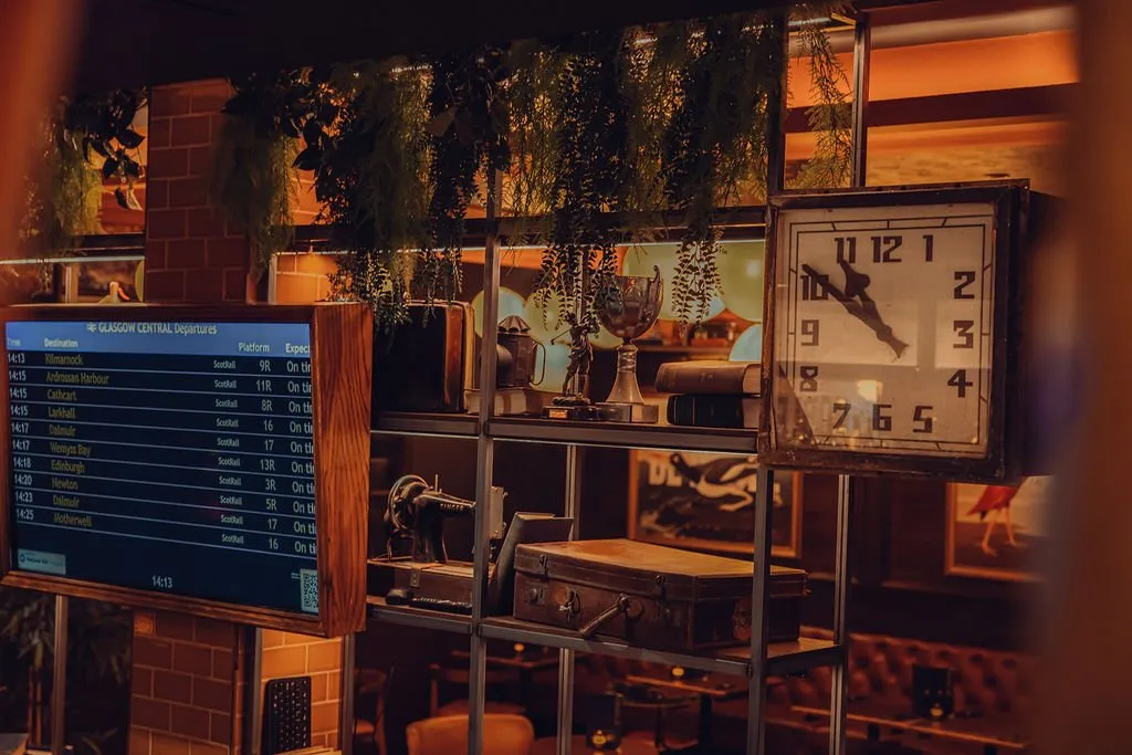A warm, vintage-inspired interior features a digital departure board, antique clock, and shelves filled with travel and industrial memorabilia, set against a brick wall.