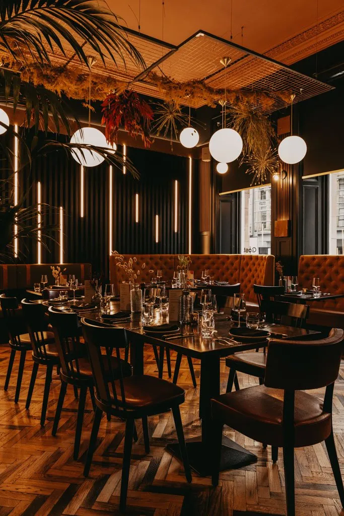 This sophisticated restaurant interior features a warm, inviting ambiance, dominated by rich, dark tones and a striking interplay of light. Dark fluted wall panels are accented by vertical amber LED strips, complementing the deep terracotta tufted banquettes and dark wood tables and chairs. Overhead, spherical pendant lights and decorative metal grids adorned with dried foliage create a lush, intimate atmosphere, all grounded by a classic herringbone wood floor.
