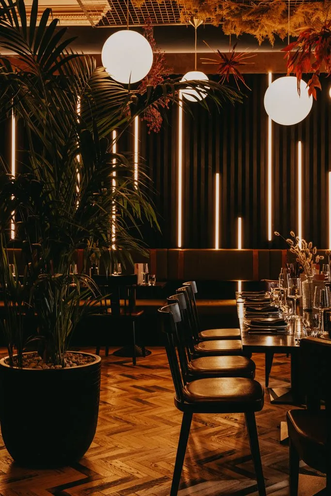 This sophisticated restaurant interior features a warm, intimate ambiance, achieved through dim, strategically placed lighting. A herringbone wood floor grounds the space, leading to a long dining table set with dark tableware and flanked by dark wood chairs. The back wall is a striking composition of dark vertical slats interspersed with glowing amber LED strips, complemented by a plush, dark upholstered banquette. Overhead, white spherical pendant lights and artful dried foliage installations add a touch of organic modernism, while a large potted palm in the foreground enhances the lush, inviting atmosphere.