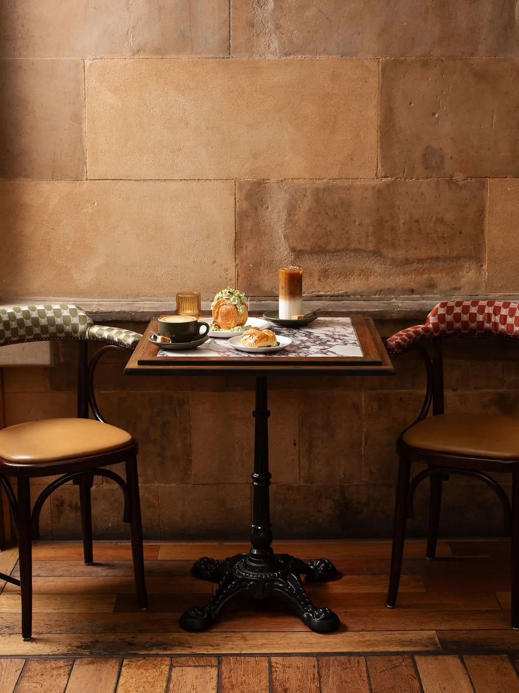 This intimate cafe setting features a square bistro table with a veined marble top and dark wood trim, supported by an ornate black cast iron pedestal base. Two classic bentwood chairs with tan leather seats flank the table, each adorned with a distinct checkerboard upholstered backrest – one green and white, the other red and white. The backdrop is a rustic wall of large, warm-toned natural stone blocks, complemented by distressed wide-plank wooden flooring, all bathed in soft, warm lighting that creates a cozy and inviting mood.