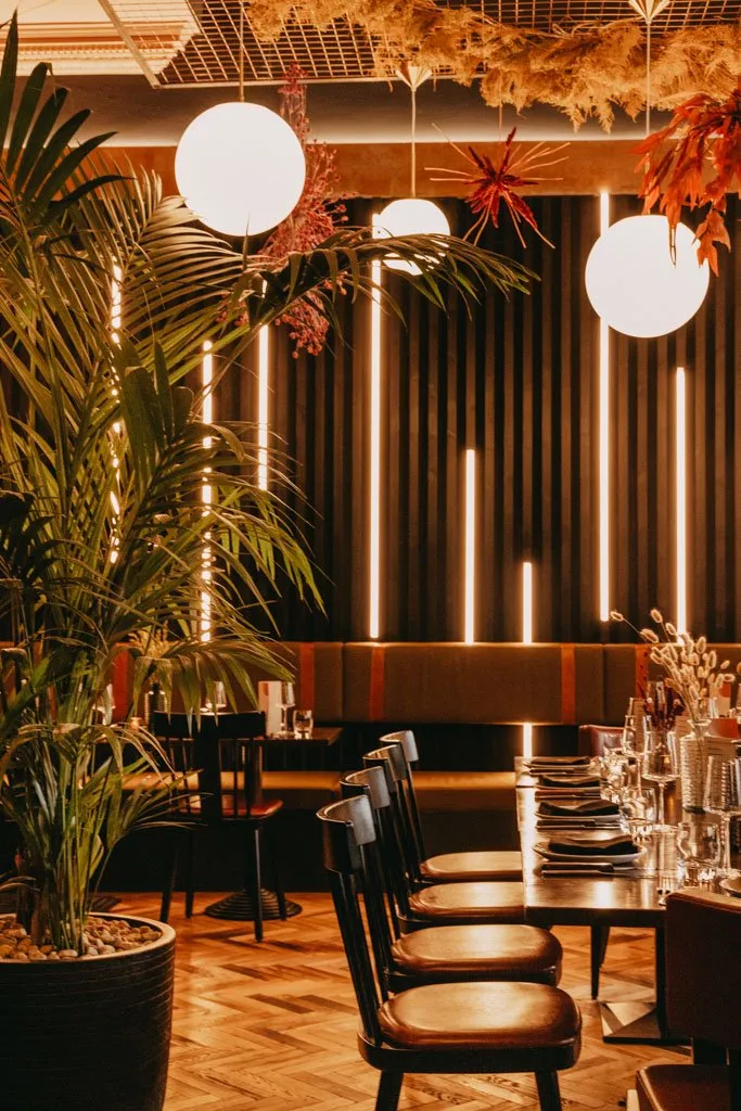 This inviting restaurant interior features a sophisticated dining area with a long olive-green banquette against a dark, fluted feature wall. Vertical LED strip lights punctuate the wall, creating a warm, rhythmic glow that complements the large spherical pendant lights and the dried autumnal foliage suspended from a metal grid ceiling. Dark wood dining chairs with rich brown leather seats and matching tables are arranged on a warm herringbone wood floor, creating an intimate and modern atmosphere.