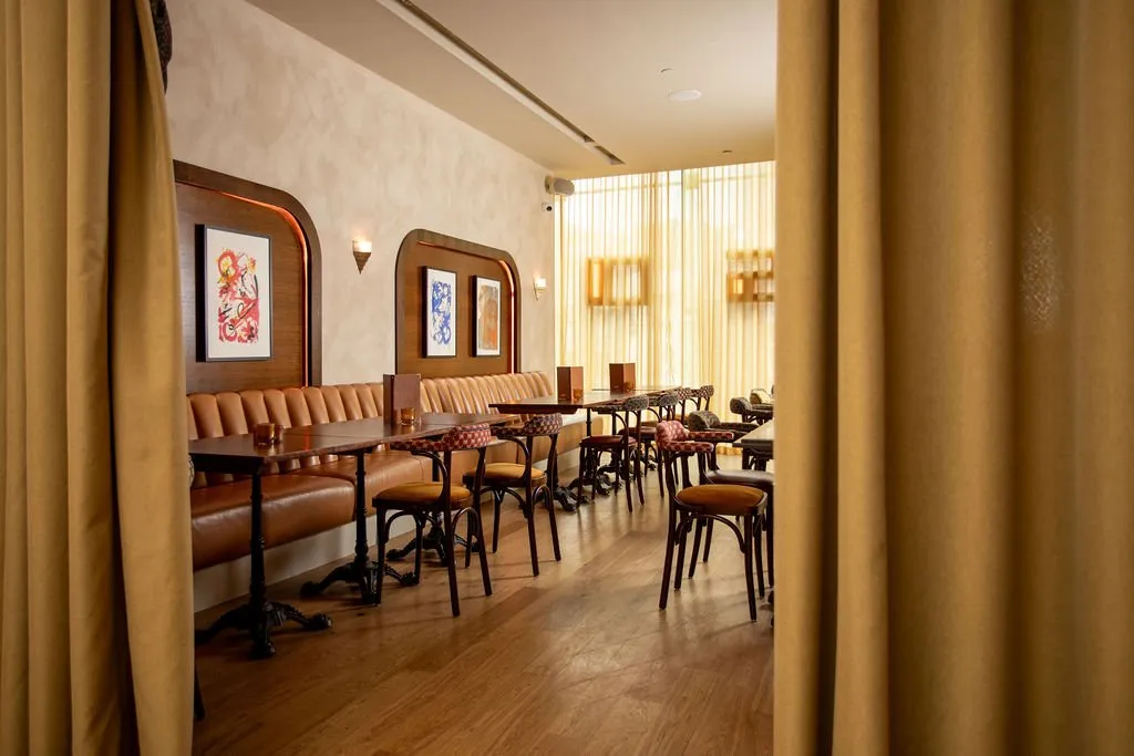 This inviting restaurant interior features a long, tan leather banquette set against a textured, light plaster wall, framed by elegant dark wood paneling with integrated warm backlighting. Individual dark wood tables with cast iron bases and bentwood chairs, some with patterned upholstery, line the space, leading towards a window wall draped in sheer, warm-toned curtains that diffuse natural light. The overall mood is warm and sophisticated, enhanced by decorative wall sconces and rich material textures, creating an intimate dining experience.