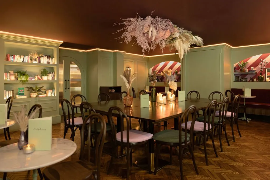 A warm and inviting private dining room features sage green walls, dark wood furniture, and a dramatic dried floral ceiling installation, creating an intimate and eclectic atmosphere.