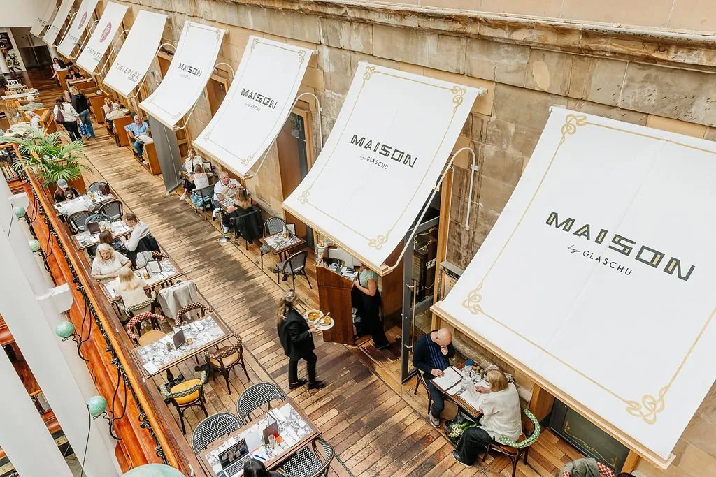 This vibrant interior shot captures a bustling multi-level dining hall, possibly a converted market or arcade, featuring a series of charming cafe-style shopfronts with white canvas awnings. Guests are seated at an eclectic mix of bistro chairs and patterned upholstered seating around small, ornate marble-topped tables, all set on a warm, distressed wooden floor. The space exudes a lively, European brasserie atmosphere, enhanced by natural light filtering down and the grand stone architecture.